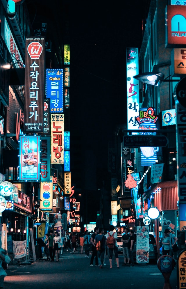 Neon lights line the streets of Seoul during a pitch black night while people gather around different stores on a footpath