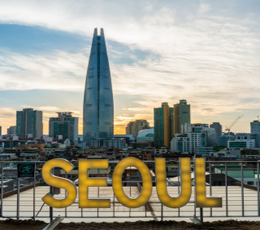 A railing with the word SEOUL written in neon letters is contrasted by a view of the cities skyline while the sun either rising or setting